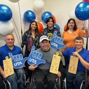 3 people in wheelchairs holding Mavup and award with three adults in the back. Plastic balloons in the background.